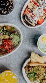 An assortment of colorful food items arranged in bowls on a textured surface. Each dish features fresh ingredients such as fruits, vegetables, and grains with vibrant toppings. Beverages accompany the dishes, enhancing the display with various colors.