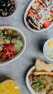 An assortment of colorful food items arranged in bowls on a textured surface. Each dish features fresh ingredients such as fruits, vegetables, and grains with vibrant toppings. Beverages accompany the dishes, enhancing the display with various colors.