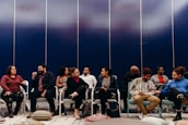 A group of people seated in a semi-circle appear to be engaged in conversation. They are sitting on chairs in a room with a dark blue paneled wall. Colorful floor cushions and a patterned rug are visible, adding a cozy feel to the setting.