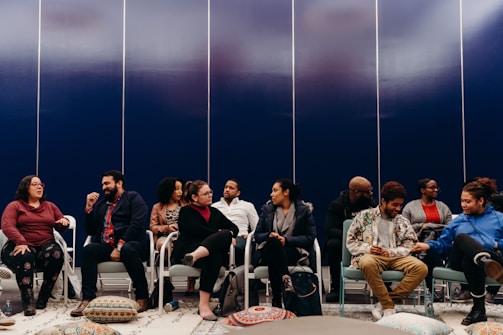 A group of people seated in a semi-circle appear to be engaged in conversation. They are sitting on chairs in a room with a dark blue paneled wall. Colorful floor cushions and a patterned rug are visible, adding a cozy feel to the setting.