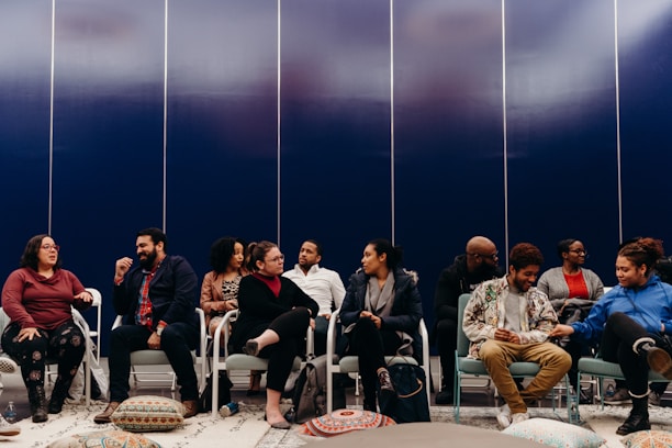 A group of people seated in a semi-circle appear to be engaged in conversation. They are sitting on chairs in a room with a dark blue paneled wall. Colorful floor cushions and a patterned rug are visible, adding a cozy feel to the setting.