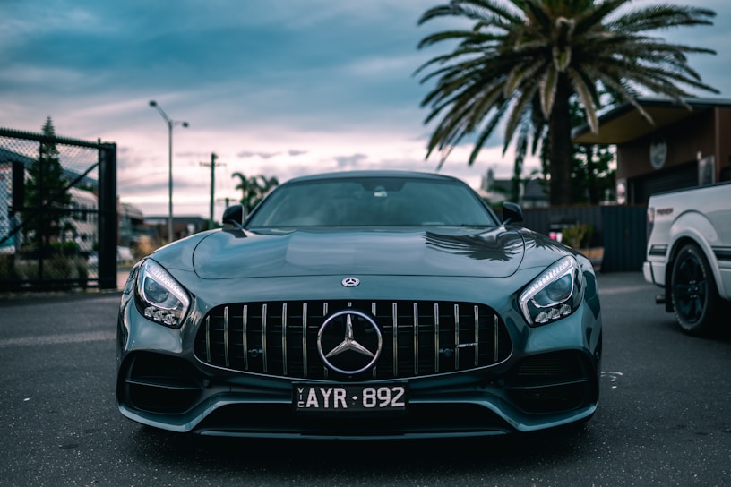 A sleek, dark green Mercedes-Benz car is prominently displayed in the center of the image. The car is parked on a street with a palm tree and some buildings in the background. The sky appears to be overcast, creating a moody atmosphere. The car's headlights are on, and the front grille and logo are clearly visible.