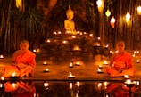 Two young monks in orange robes are seated cross-legged in meditation beside a small pond, surrounded by numerous lit candles. A Buddha statue is prominently displayed on a raised platform behind them, set against a backdrop of bamboo and hanging lanterns.