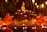 Two young monks in orange robes are seated cross-legged in meditation beside a small pond, surrounded by numerous lit candles. A Buddha statue is prominently displayed on a raised platform behind them, set against a backdrop of bamboo and hanging lanterns.