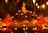 Two young monks in orange robes are seated cross-legged in meditation beside a small pond, surrounded by numerous lit candles. A Buddha statue is prominently displayed on a raised platform behind them, set against a backdrop of bamboo and hanging lanterns.