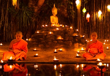 Two young monks in orange robes are seated cross-legged in meditation beside a small pond, surrounded by numerous lit candles. A Buddha statue is prominently displayed on a raised platform behind them, set against a backdrop of bamboo and hanging lanterns.