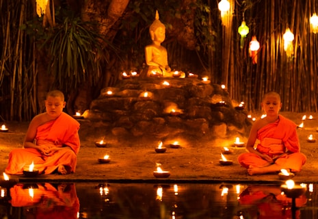 Two young monks in orange robes are seated cross-legged in meditation beside a small pond, surrounded by numerous lit candles. A Buddha statue is prominently displayed on a raised platform behind them, set against a backdrop of bamboo and hanging lanterns.