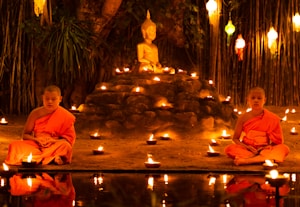 Two young monks in orange robes are seated cross-legged in meditation beside a small pond, surrounded by numerous lit candles. A Buddha statue is prominently displayed on a raised platform behind them, set against a backdrop of bamboo and hanging lanterns.