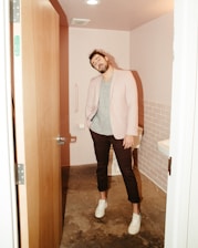 A man stands in a small bathroom with pink-tiled walls. He is casually dressed in a light blazer over a striped t-shirt, dark pants, and white sneakers. The bathroom has a partly open wooden door, a toilet, a grab bar, and a sink partially visible on the right side.
