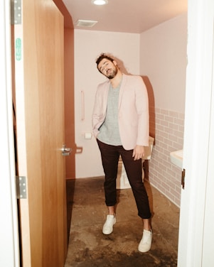 A man stands in a small bathroom with pink-tiled walls. He is casually dressed in a light blazer over a striped t-shirt, dark pants, and white sneakers. The bathroom has a partly open wooden door, a toilet, a grab bar, and a sink partially visible on the right side.