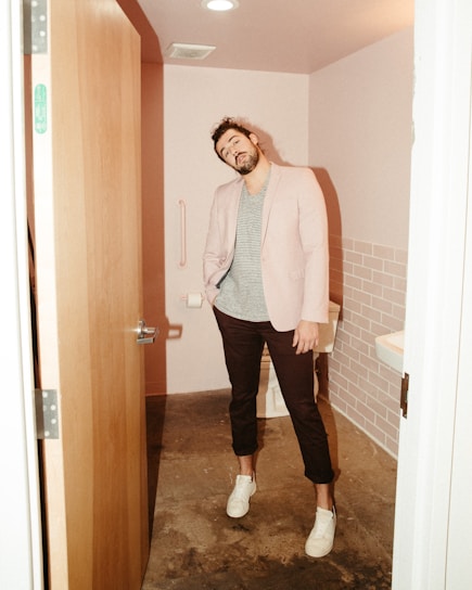 A man stands in a small bathroom with pink-tiled walls. He is casually dressed in a light blazer over a striped t-shirt, dark pants, and white sneakers. The bathroom has a partly open wooden door, a toilet, a grab bar, and a sink partially visible on the right side.