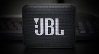 A small, portable speaker with a rectangular shape and a grille on the front displaying the JBL logo in prominent white letters. The speaker is set against a dark, blurry background, adding a sleek and modern aesthetic.