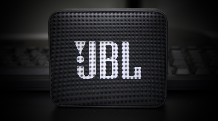 A small, portable speaker with a rectangular shape and a grille on the front displaying the JBL logo in prominent white letters. The speaker is set against a dark, blurry background, adding a sleek and modern aesthetic.