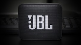 A small, portable speaker with a rectangular shape and a grille on the front displaying the JBL logo in prominent white letters. The speaker is set against a dark, blurry background, adding a sleek and modern aesthetic.