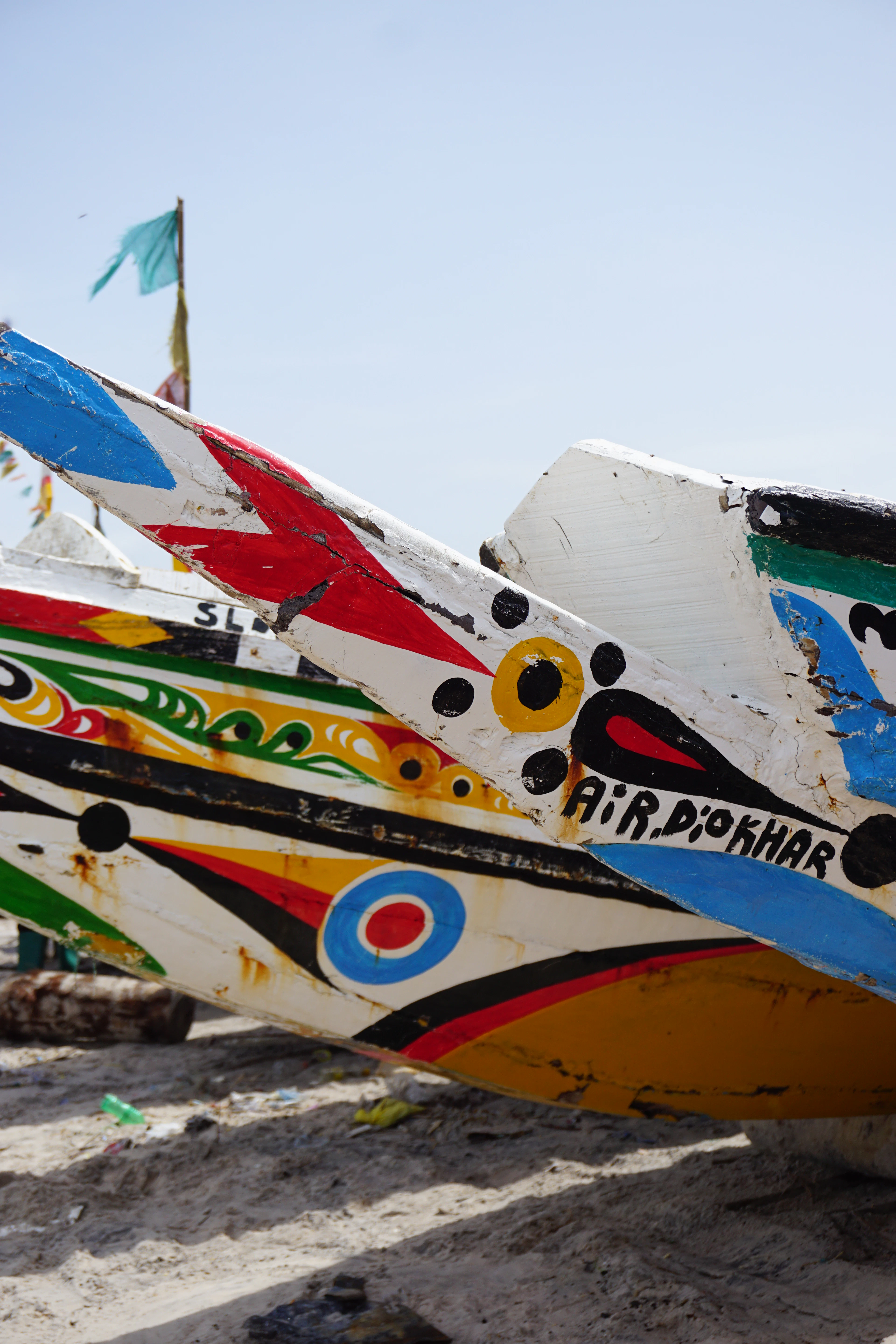 The bows of two hand-painted Senegalese fishing pirogues hauled onto a Dakar beach, their hulls covered in bold geometric motifs, eyes and lettering