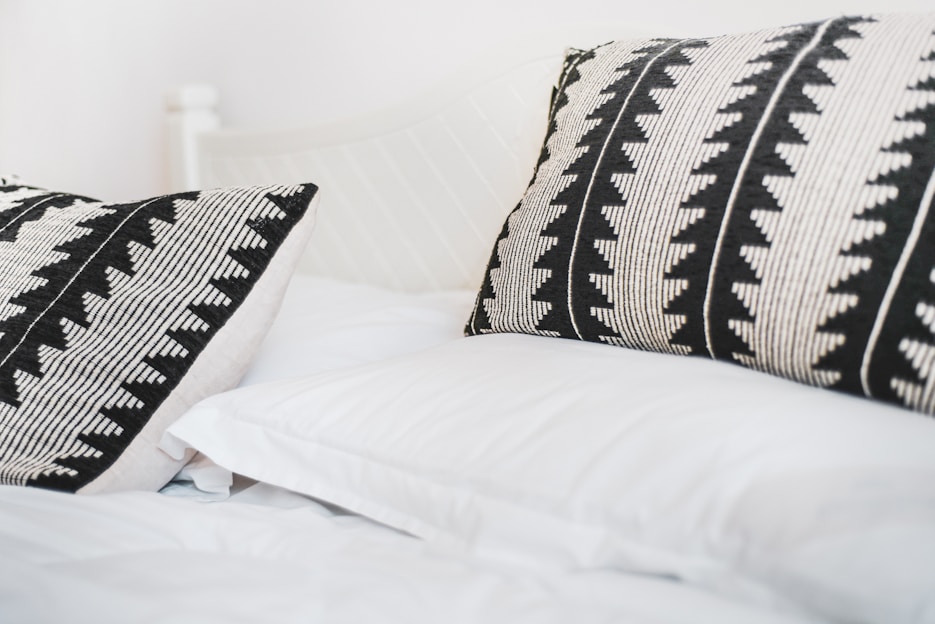 Two decorative pillows with black and white geometric patterns rest on a neatly made bed with white sheets. The overall setting is minimalist and serene, giving a sense of comfort and simplicity.