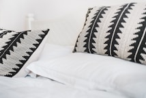 Two decorative pillows with black and white geometric patterns rest on a neatly made bed with white sheets. The overall setting is minimalist and serene, giving a sense of comfort and simplicity.