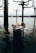 A trash bin is partially submerged in floodwaters, containing various pieces of litter such as cups and cartons. Leafless trees stand in the background with a view of a distant town across the water under an overcast sky.