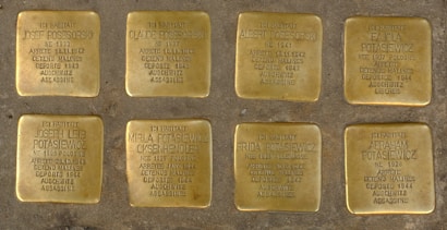 The image contains eight brass plaques embedded in the ground. Each plaque bears inscriptions commemorating victims of the Holocaust, detailing their names, years of birth, arrest dates, detention locations, deportation to Auschwitz, and their fate. The plaques are placed in a grid formation on a pavement background.