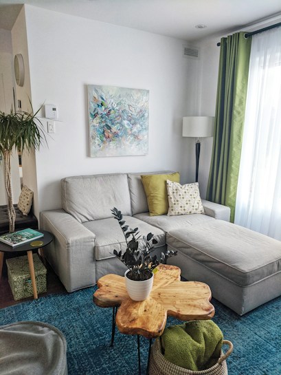 A cozy living room with a light gray sectional sofa adorned with green and patterned pillows. A botanical-themed abstract painting hangs on the white wall behind the sofa. A unique wooden coffee table, shaped like a slice of tree trunk, holds a potted plant with dark green leaves. A blue area rug covers the floor, and a wicker basket with a green blanket is tucked underneath the table. A tall lamp with a white shade stands near the corner, providing soft lighting. Light green curtains border the large window, allowing natural light to illuminate the space.
