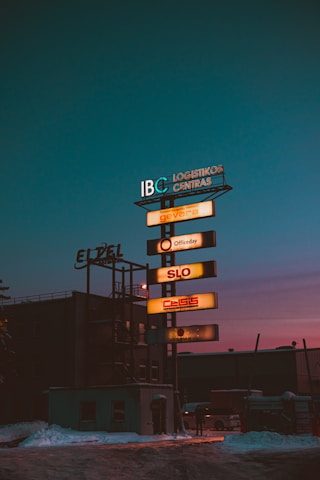 A tall signpost displays various company names in illuminated panels, set against a darkening sky with a hint of dusk colors. Snow is visible on the ground, and the surrounding buildings are dimly lit, indicating a possibly industrial or commercial area.