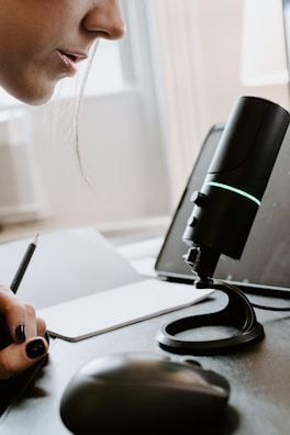A close-up view of a person speaking into a microphone, with a laptop and mouse on a desk. The person is holding a pencil and a notebook is visible. The focus is on the microphone, which is highlighted by an LED light.