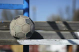 A young man sitting on a bench, looking thoughtfully at a soccer ball under a sunset sky.