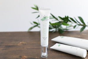 A tube of skincare cream labeled 'CRYSTALWEED High Quality CBD' is prominently displayed on a wooden surface. In the background, there are green leaves, adding a natural element to the setting. Additional tubes lie on the surface, slightly out of focus.