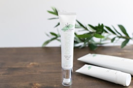 A tube of skincare cream labeled 'CRYSTALWEED High Quality CBD' is prominently displayed on a wooden surface. In the background, there are green leaves, adding a natural element to the setting. Additional tubes lie on the surface, slightly out of focus.