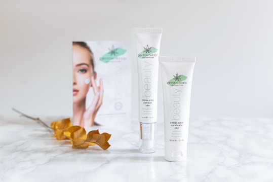 Two tubes of Crystalweed CBD beauty products are placed on a marble surface. A blurred advertisement featuring a woman's face is visible in the background. The foliage with dry, brown leaves adds a touch of nature to the scene.