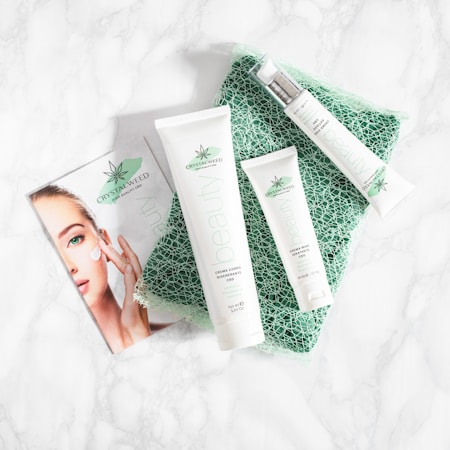 A collection of skincare products labeled with the brand 'Crystalweed', featuring a woman applying cream on her face in a promotional flyer. The products are placed on a textured green surface against a marble background.