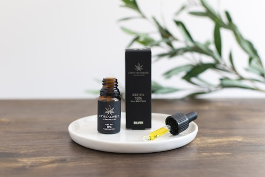 A small amber bottle labeled as CBD oil stands on a white round dish next to a dropper filled with yellow liquid. The bottle and dropper are positioned on a wooden surface, with a black box behind them. Green leaves are blurred in the background, suggesting a natural setting.
