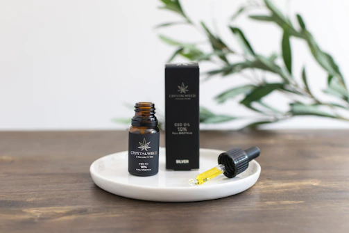 A small amber bottle labeled as CBD oil stands on a white round dish next to a dropper filled with yellow liquid. The bottle and dropper are positioned on a wooden surface, with a black box behind them. Green leaves are blurred in the background, suggesting a natural setting.