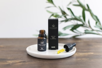 A small amber bottle labeled as CBD oil stands on a white round dish next to a dropper filled with yellow liquid. The bottle and dropper are positioned on a wooden surface, with a black box behind them. Green leaves are blurred in the background, suggesting a natural setting.