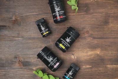A collection of black containers labeled 'Crystalweed' is arranged on a wooden surface. Each container has a different colored label indicating various products, and a small green plant is positioned near the bottom left corner.