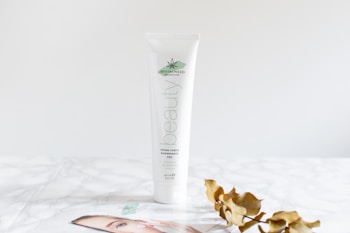 A white tube of beauty cream labeled 'Crystalweed' is standing on a white marble surface. Next to the tube is a dried branch with curled brown leaves and a partial view of a booklet or brochure featuring a close-up image of an eye. The background is a soft, neutral tone.