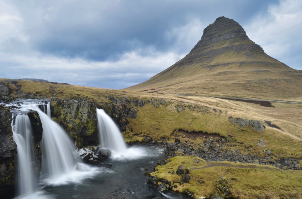 water falls near brown mountain under white clouds and blue sky during daytime