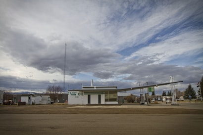 An old-fashioned gas station with a large sign reading 'VAN OIL CO.' in green letters. The structure is situated under a cloudy sky with patches of blue visible. The setting appears quiet and somewhat deserted, with no vehicles or people present. There are some bare trees and a few buildings in the background.