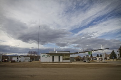 An old-fashioned gas station with a large sign reading 'VAN OIL CO.' in green letters. The structure is situated under a cloudy sky with patches of blue visible. The setting appears quiet and somewhat deserted, with no vehicles or people present. There are some bare trees and a few buildings in the background.