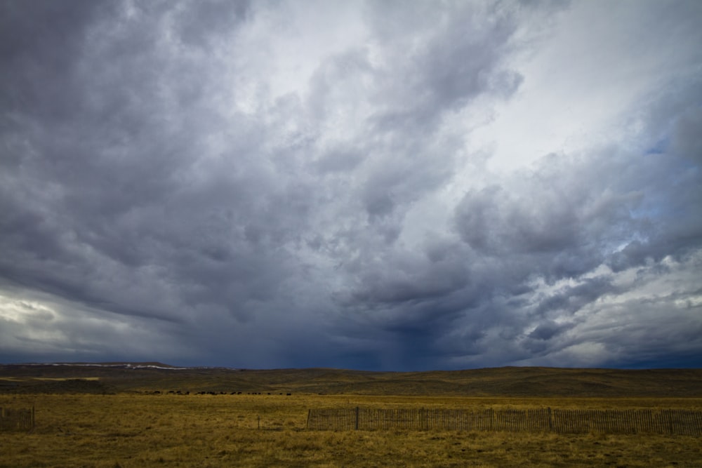 a field with a fence in the foreground and a cloudy sky in the background