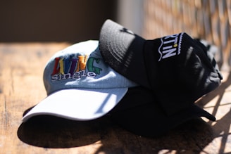 A close-up of two caps stacked on a wooden surface, with one cap displaying the words 'Living Single' in colorful embroidery. The caps are well-lit by natural sunlight, casting shadows on the surface.