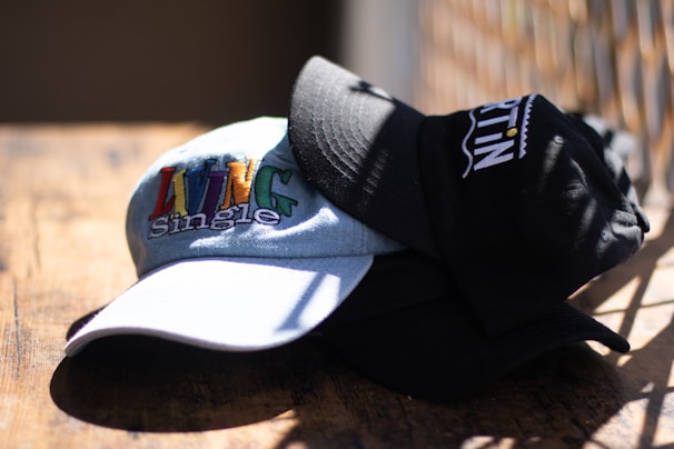 A close-up of two caps stacked on a wooden surface, with one cap displaying the words 'Living Single' in colorful embroidery. The caps are well-lit by natural sunlight, casting shadows on the surface.