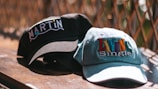 Two baseball caps are placed on a wooden surface with a wire fence in the background. One cap is black with the word 'MARTIN' embroidered in white and colored letters. The other cap is light denim with the phrase 'LIVIN Single' embroidered in colorful letters.
