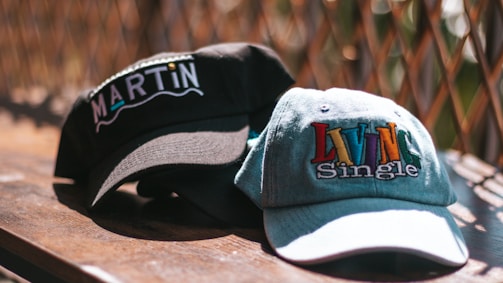 Two baseball caps are placed on a wooden surface with a wire fence in the background. One cap is black with the word 'MARTIN' embroidered in white and colored letters. The other cap is light denim with the phrase 'LIVIN Single' embroidered in colorful letters.