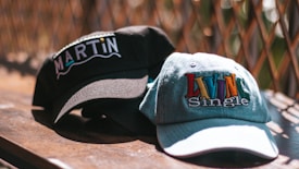 Two baseball caps are placed on a wooden surface with a wire fence in the background. One cap is black with the word 'MARTIN' embroidered in white and colored letters. The other cap is light denim with the phrase 'LIVIN Single' embroidered in colorful letters.