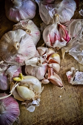 Several heads and cloves of garlic are scattered across a wooden surface. The garlic has a natural, rustic appearance with hues of purple and white. Some of the cloves are partially peeled, revealing their smooth inner texture.