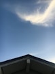 The image features a triangular rooftop against a vast blue sky with a singular cloud formation. The roof's peak creates a distinct point at the bottom of the image, contrasting with the soft, wispy clouds above. The sky is clear with a smooth gradient transitioning from a deeper blue at the top to a lighter shade near the horizon.
