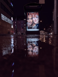 A nighttime city scene featuring a bus stop with illuminated advertisement displays. The reflections of the advertisements are visible on the wet pavement. Surrounding buildings are dimly lit, indicating a quiet urban environment.