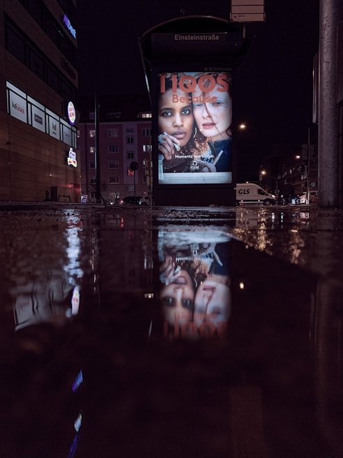 A nighttime city scene featuring a bus stop with illuminated advertisement displays. The reflections of the advertisements are visible on the wet pavement. Surrounding buildings are dimly lit, indicating a quiet urban environment.