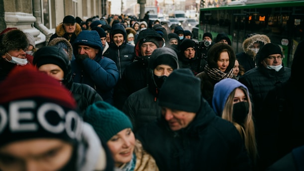 A large group of people, mostly dressed in winter clothing with hats and masks, are crowded together outdoors. They appear to be in a public space, possibly waiting in line. The background shows elements of an urban setting, including a bus in motion. The crowd's expressions are varied, with some people smiling and others appearing neutral or focused.